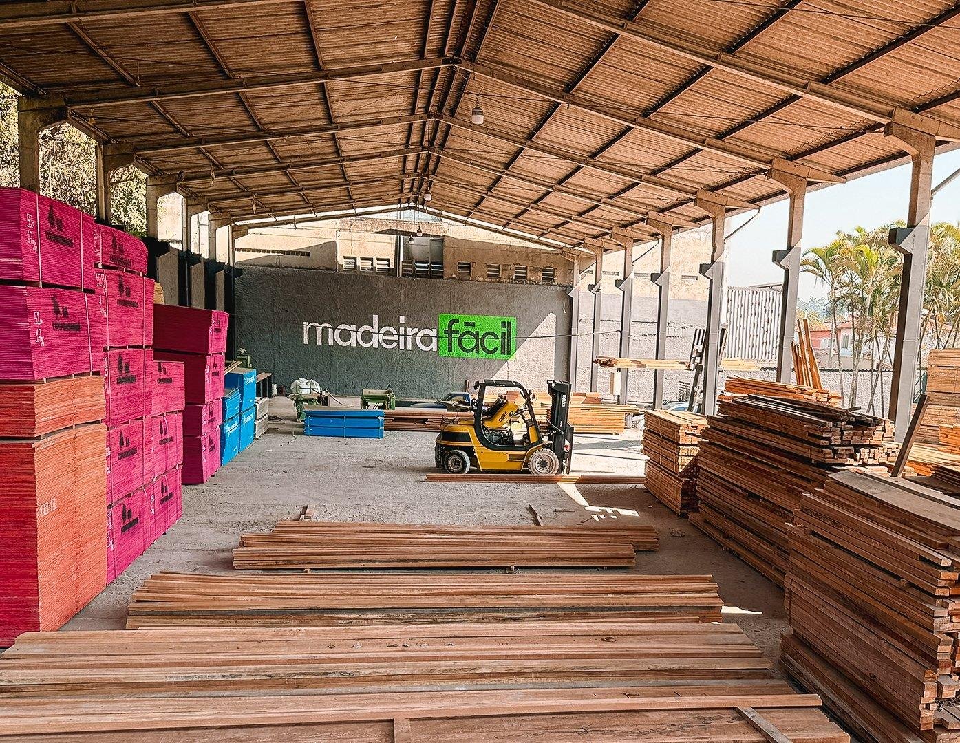 Visão ampla do interior de uma madeireira organizada com empilhadeiras e estoques de chapas e tábuas.