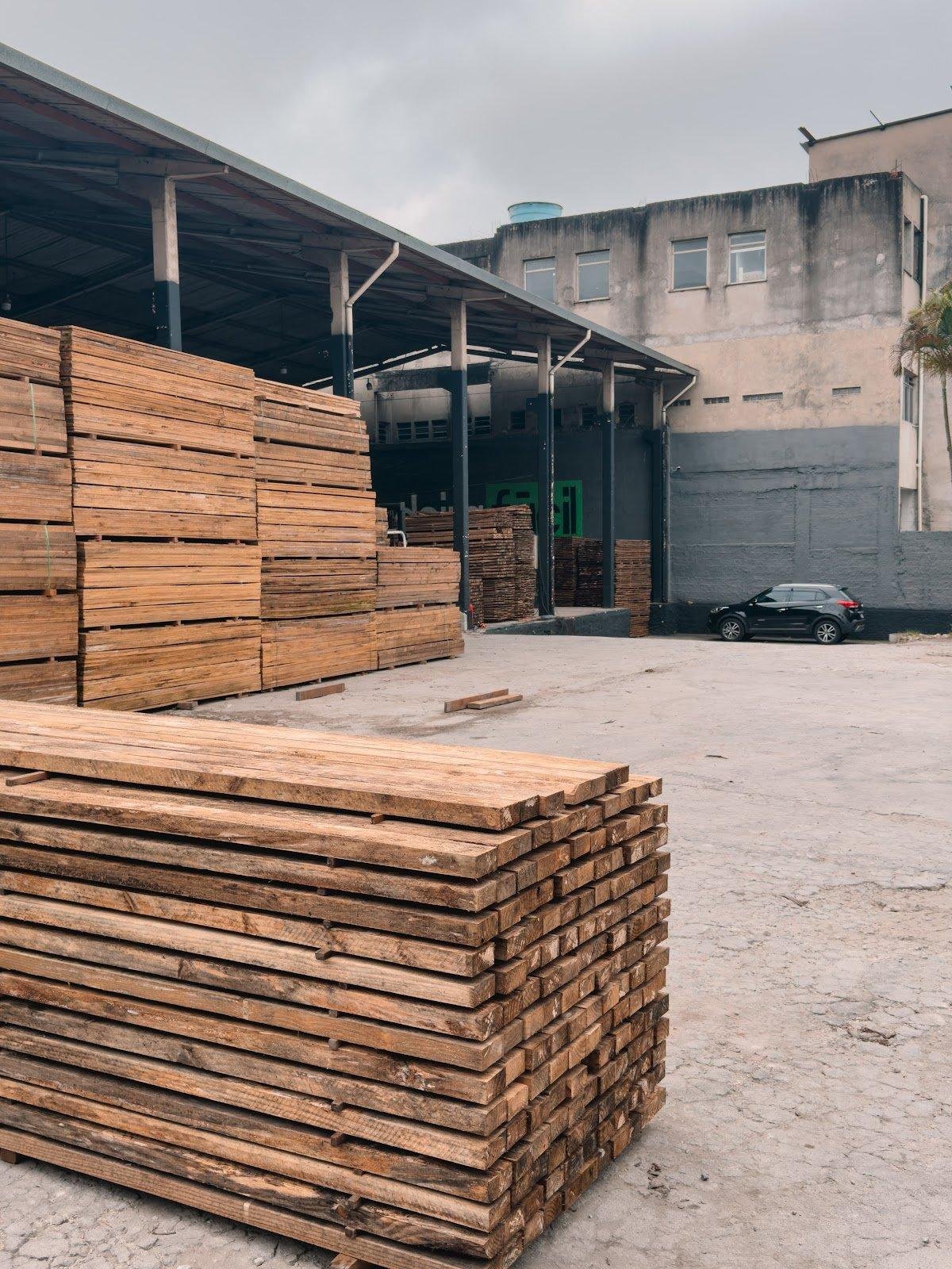 Pátio externo com grandes pilhas de madeira serrada e estrutura de galpão.